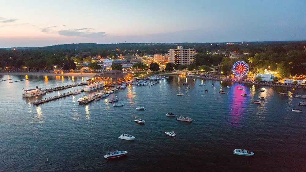 Aerial view of the Lake Geneva waterfront at dusk, with boats in the harbor, piers lit along the shore and a Ferris wheel glowing on the beach.